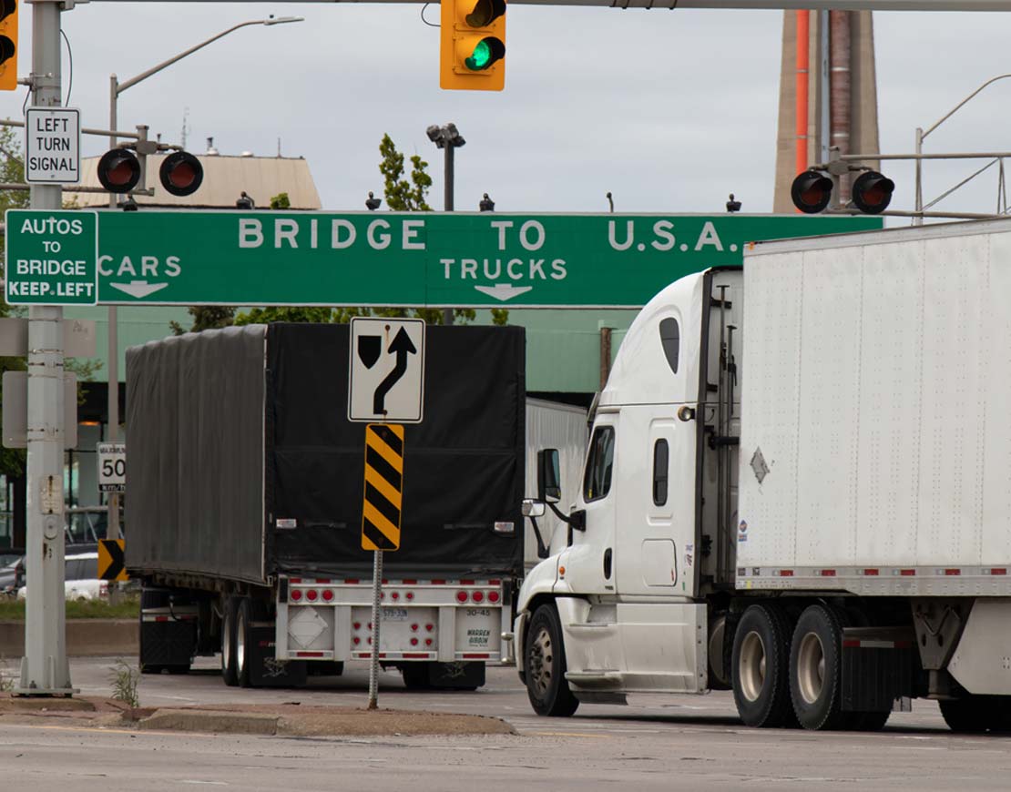 Cargo trucks passing under a sign with bridge to U.S.A. – transborder services
